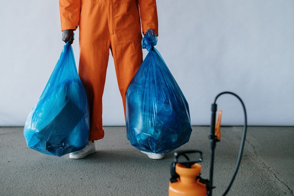 Close-up of a person wearing orange overalls and white gloves, holding a blue plastic rubbish bag with gathered material inside, standing outdoors on a concrete surface with a blurred background. The